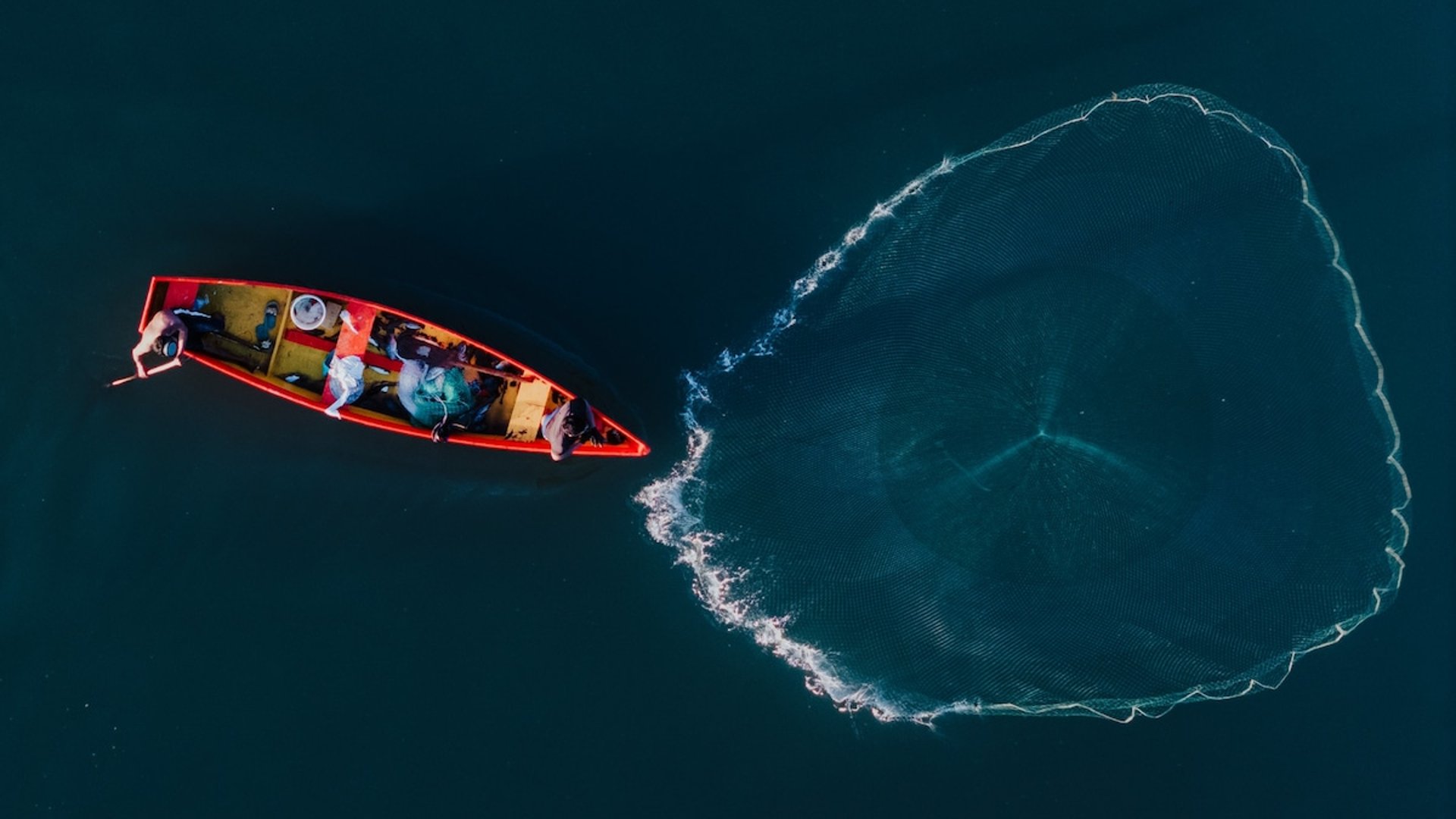Vista aérea de pescadores en lancha roja lanzando red circular sobre aguas profundas y tranquilas