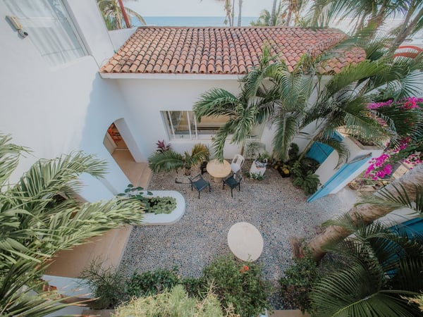 Vista aérea del patio interior empedrado con palmeras, buganvilias y villa blanca de techo de teja, mar al fondo