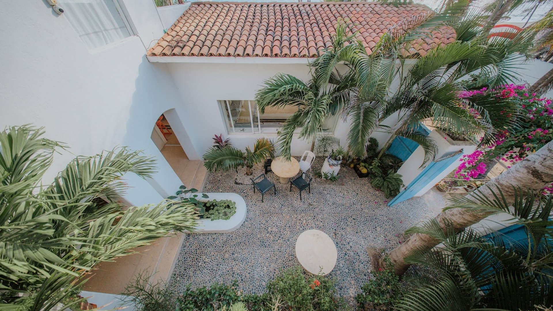 Vista aérea del patio interior empedrado con palmeras, buganvilias y villa blanca de techo de teja, mar al fondo