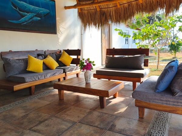 Sala exterior bajo palapa con muebles de madera maciza, cojines grises y amarillos, y cuadro de ballenas en la pared