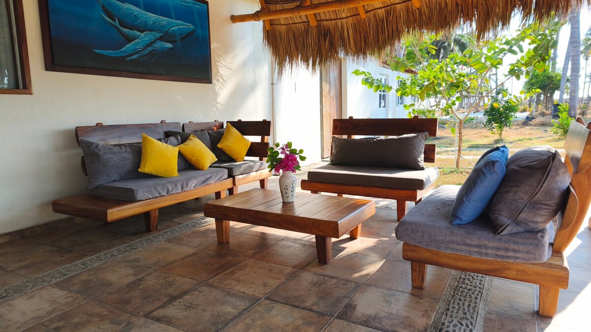 Sala exterior bajo palapa con muebles de madera maciza, cojines grises y amarillos, y cuadro de ballenas en la pared