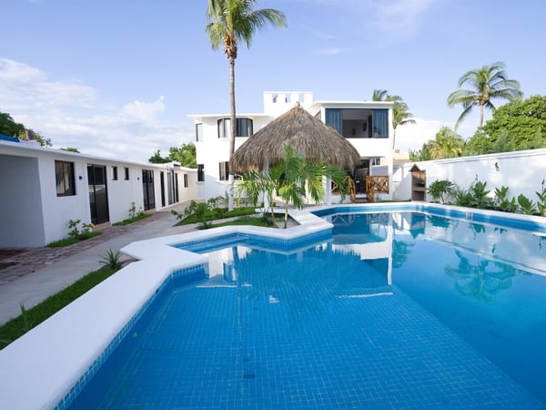 Amplia alberca azul con palapa central y villa de dos pisos al fondo, rodeada de palmeras y jardín en día soleado
