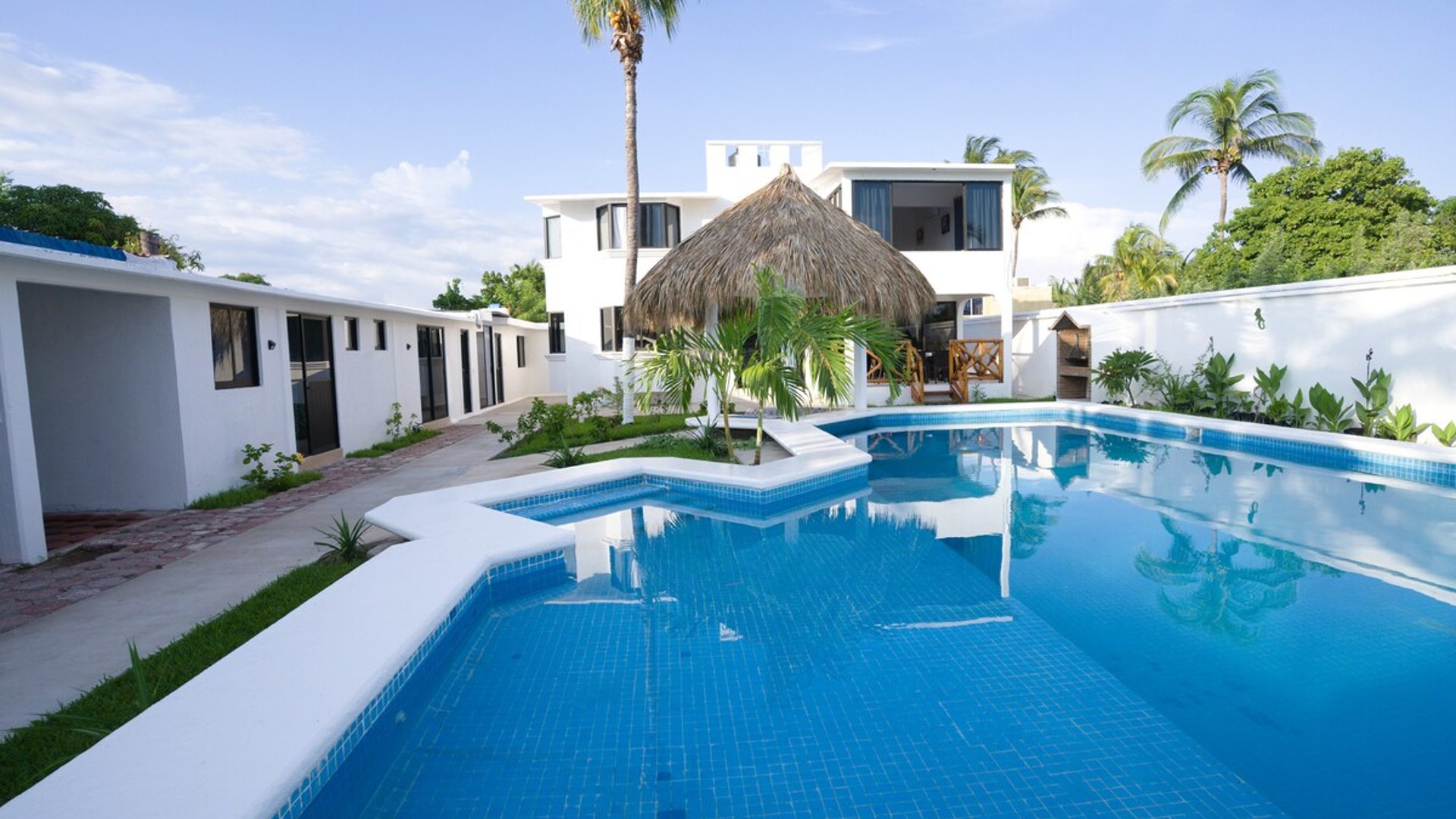 Amplia alberca azul con palapa central y villa de dos pisos al fondo, rodeada de palmeras y jardín en día soleado