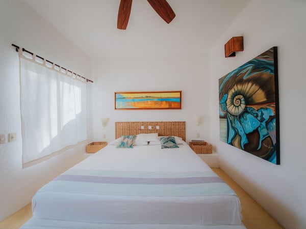 Recámara blanca con cama doble, cabecera de mimbre, ventilador de madera y cuadros de arte coloridos