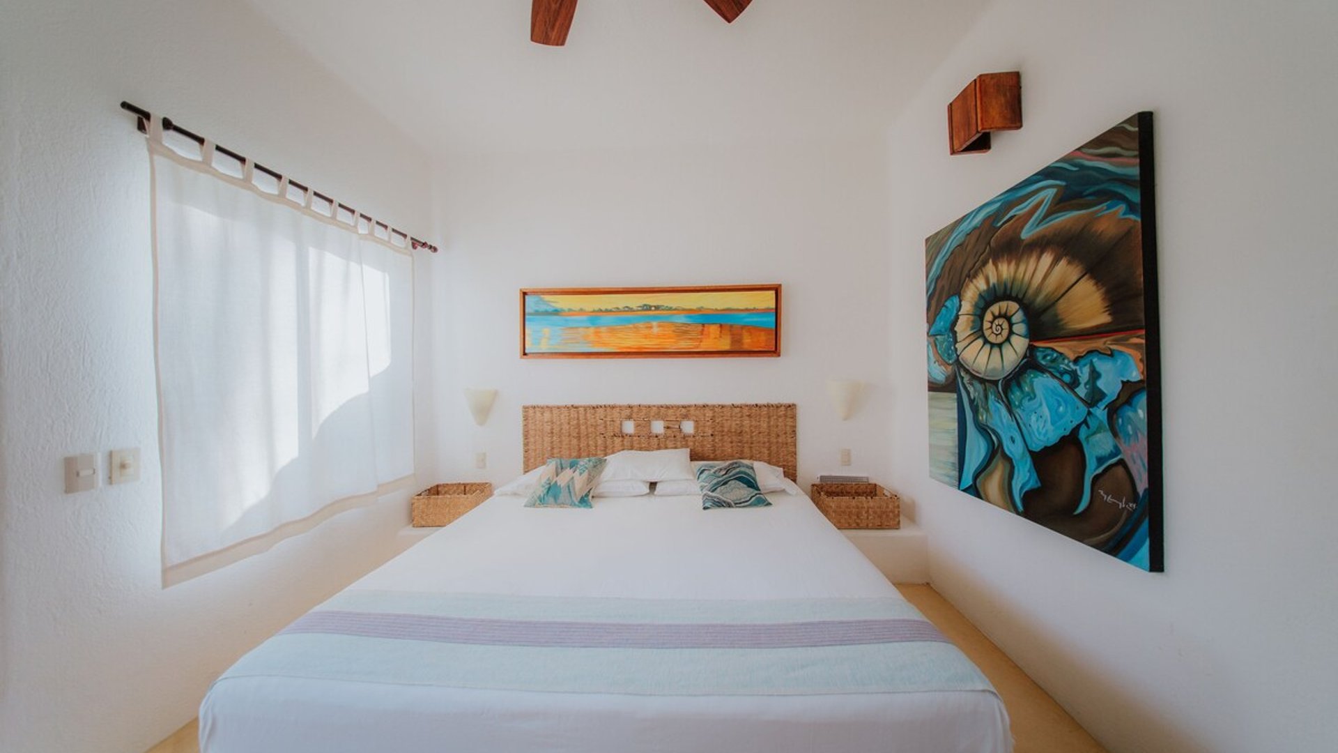 Recámara blanca con cama doble, cabecera de mimbre, ventilador de madera y cuadros de arte coloridos