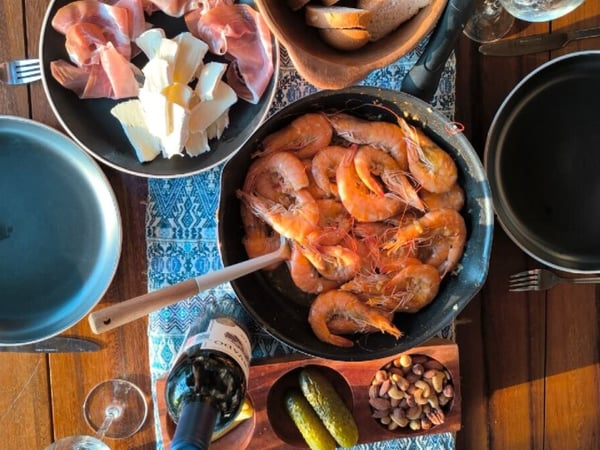 Mesa de madera con camarones al ajillo en sartén, tabla de embutidos y quesos, vino tinto y botanas variadas