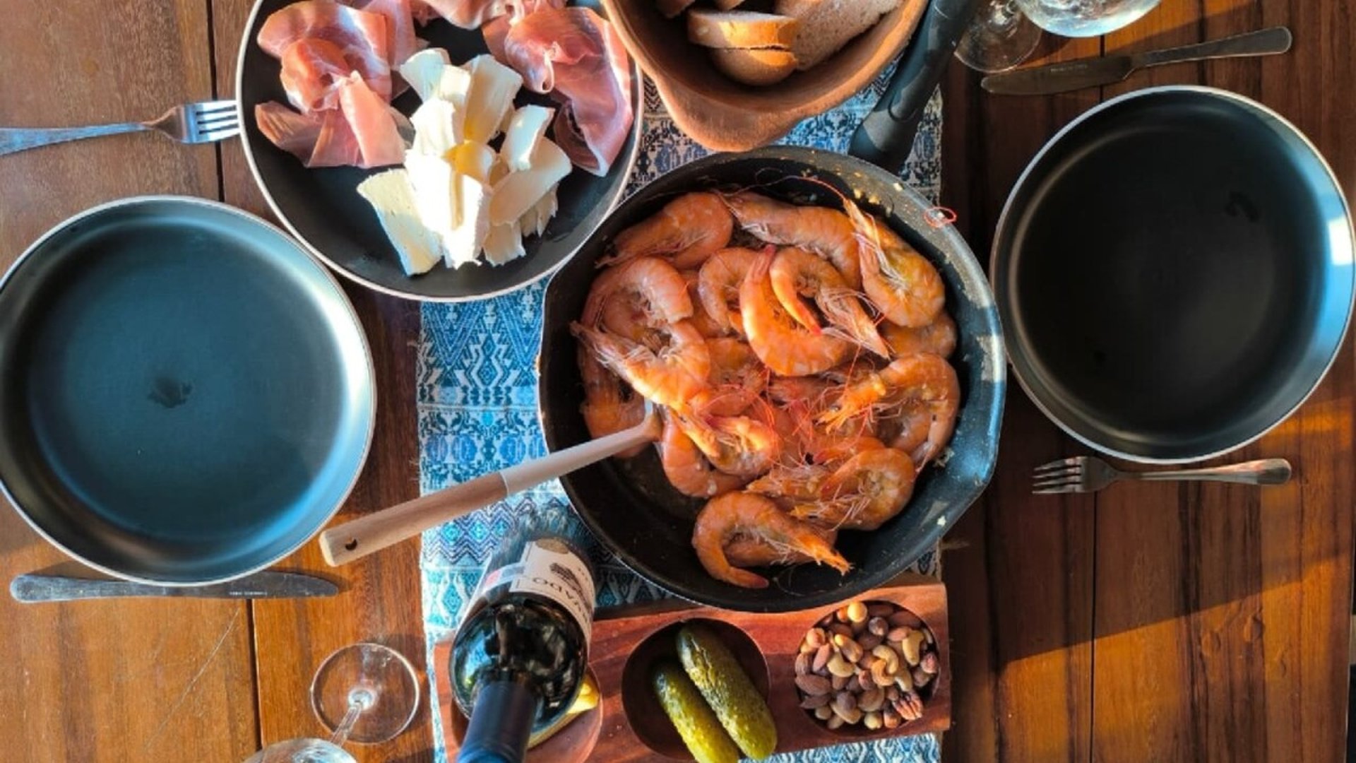 Mesa de madera con camarones al ajillo en sartén, tabla de embutidos y quesos, vino tinto y botanas variadas