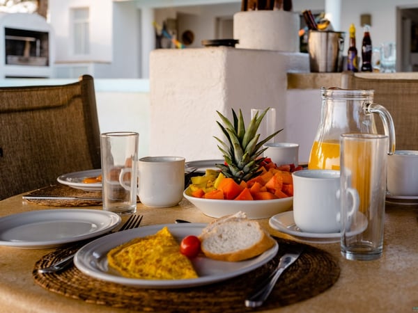 Mesa de desayuno exterior con omelette, frutas tropicales, jugo de naranja y café, con cocina al fondo