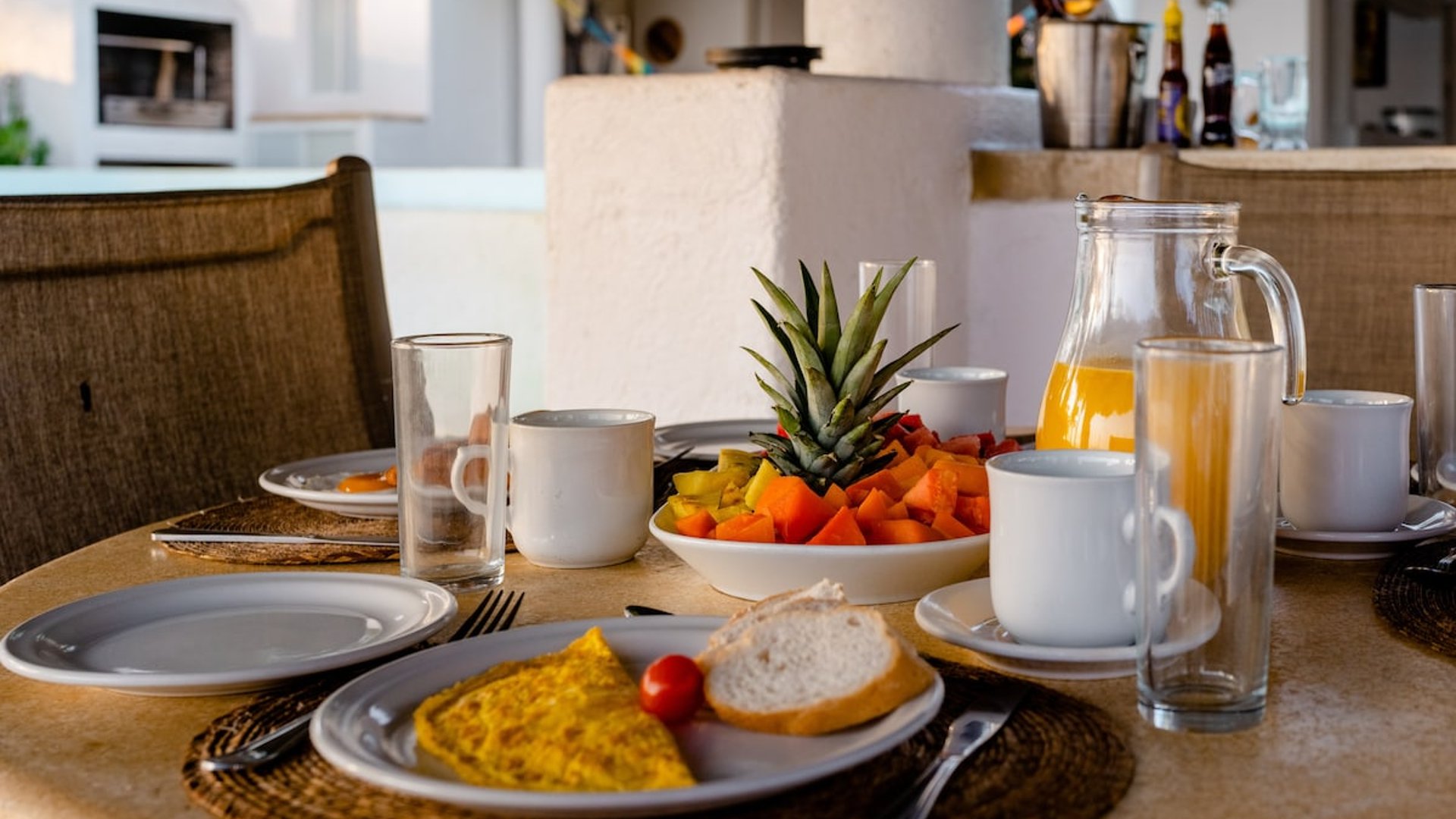 Mesa de desayuno exterior con omelette, frutas tropicales, jugo de naranja y café, con cocina al fondo