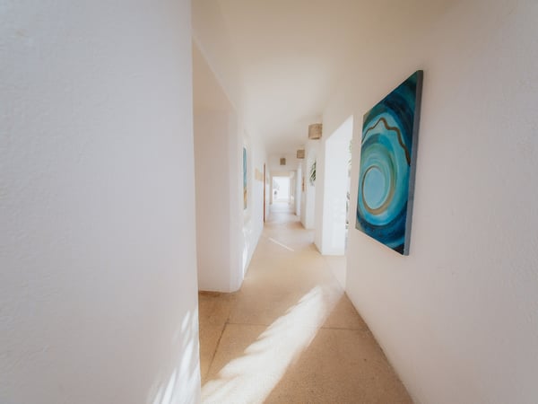 Pasillo interior luminoso con paredes blancas y cuadros de tonos turquesa, bañado por luz natural
