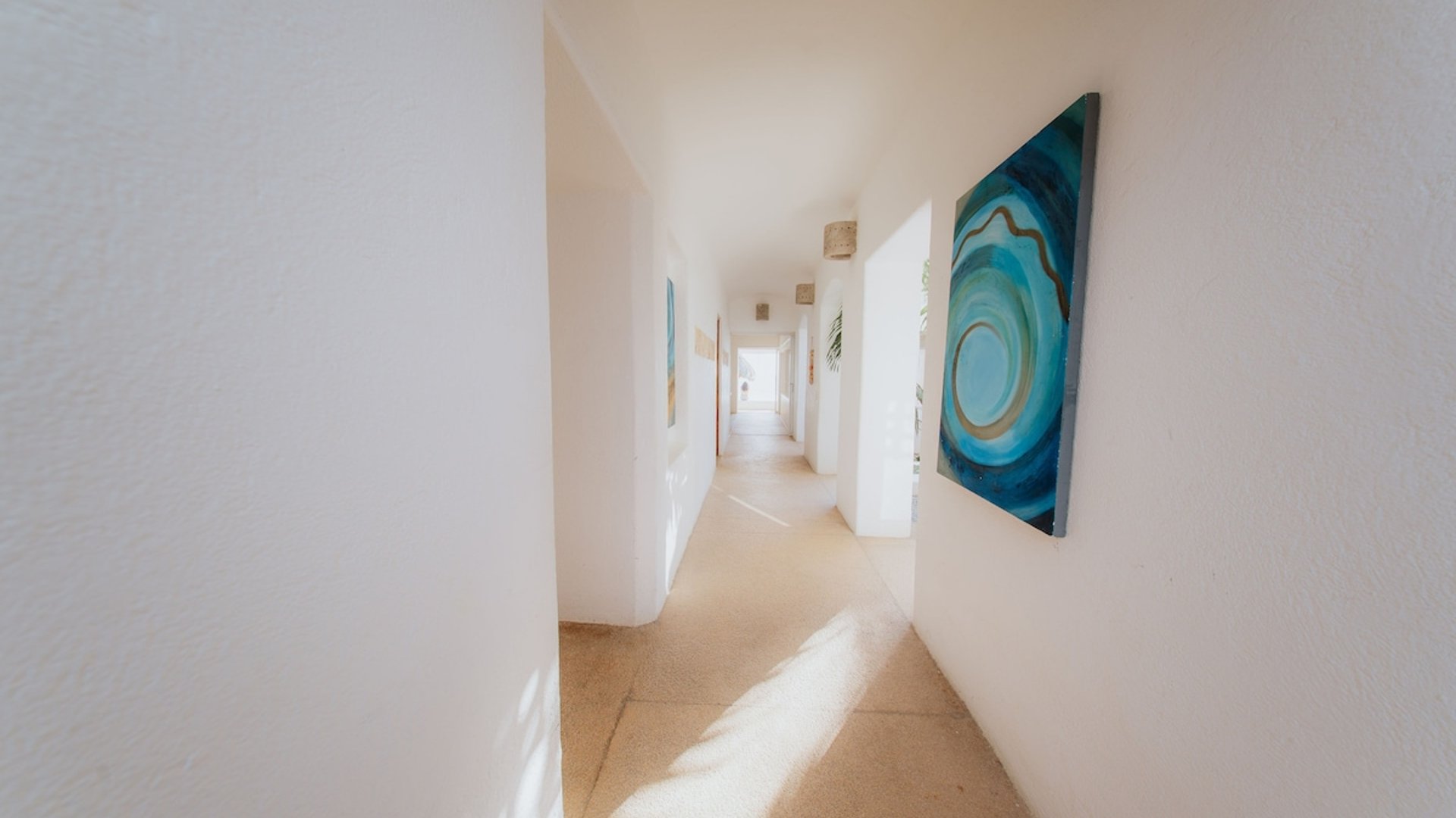 Pasillo interior luminoso con paredes blancas y cuadros de tonos turquesa, bañado por luz natural