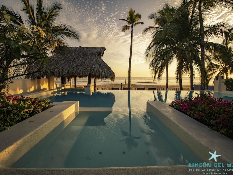 Alberca infinity y jacuzzi con palapa frente al mar al atardecer, rodeados de palmeras y buganvilias