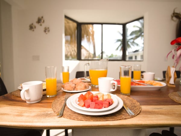 Mesa de comedor interior servida con frutas, jugo de naranja y pan, con vista a palmeras por ventana