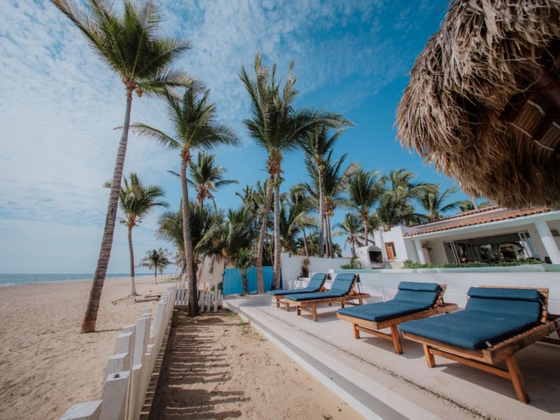 Terraza de playa con camastros azules bajo palmas de coco, palapa y villa blanca al fondo con vista al mar