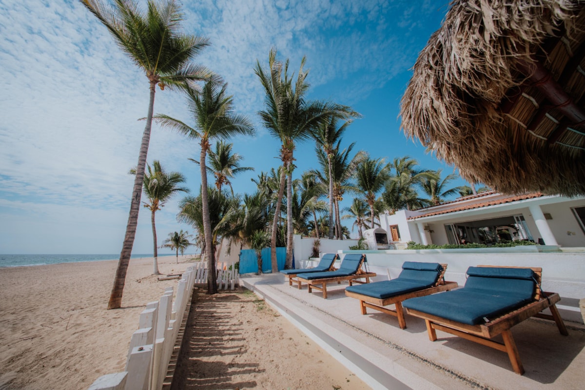 Terraza de playa con camastros azules bajo palmas y palapa de Rincón del Mar