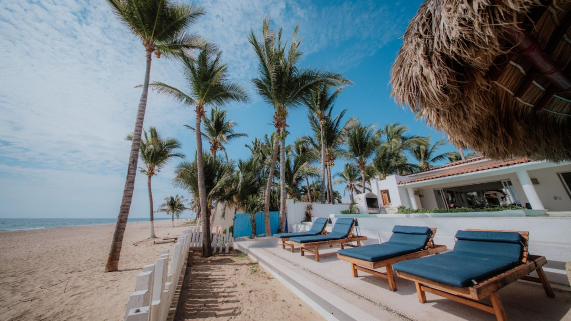Terraza de playa con camastros azules bajo palmas de coco, palapa y villa blanca al fondo con vista al mar