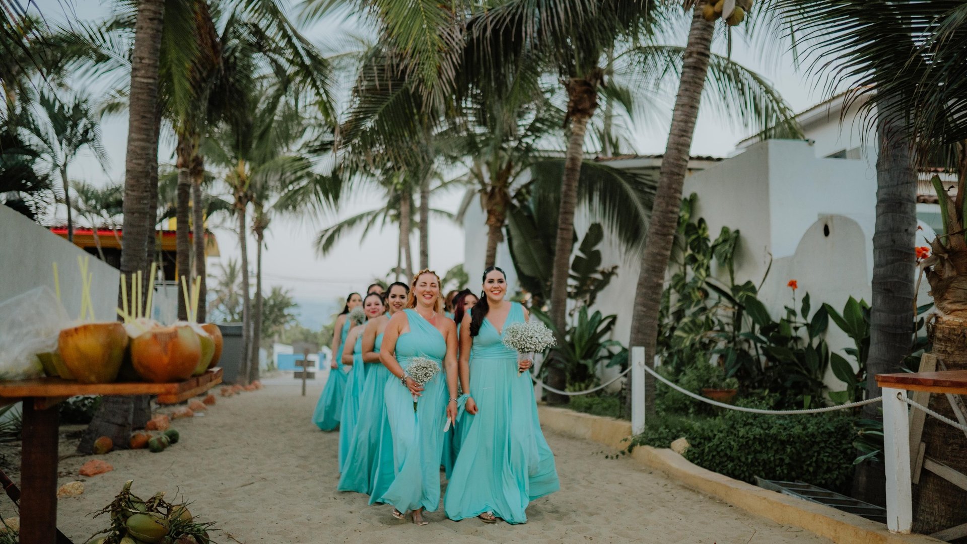 Cortejo de damas con vestidos turquesa caminando por sendero entre palmeras hacia vista al mar