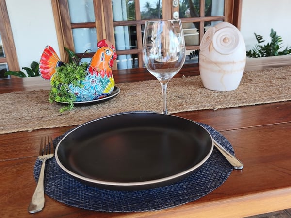 Mesa de madera con lugar individual montado: plato negro mate, copa de vino y figura de gallo talavera como decoración