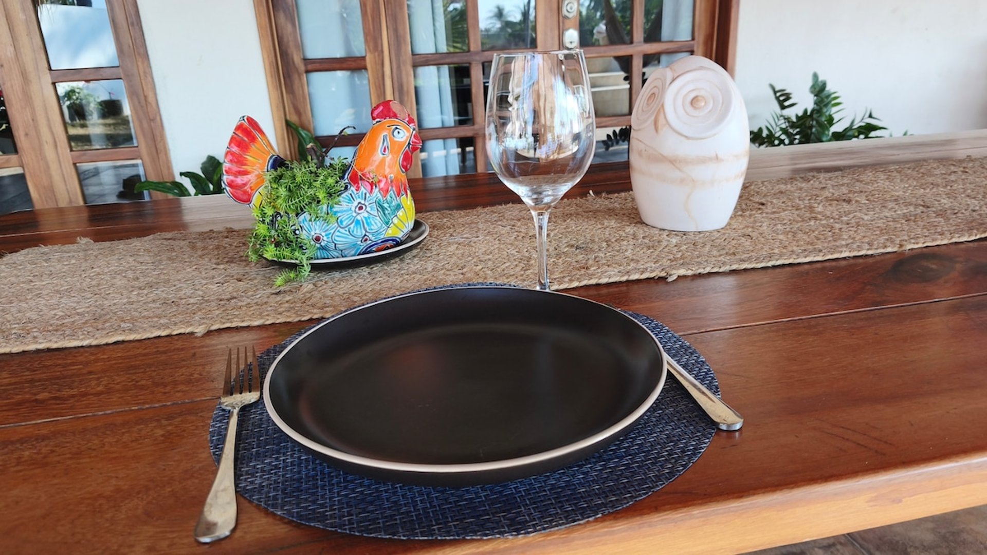 Mesa de madera con lugar individual montado: plato negro mate, copa de vino y figura de gallo talavera como decoración