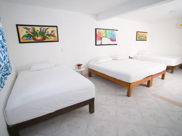 Recámara amplia con tres camas de madera, ropa de cama blanca y cuadros coloridos en paredes blancas