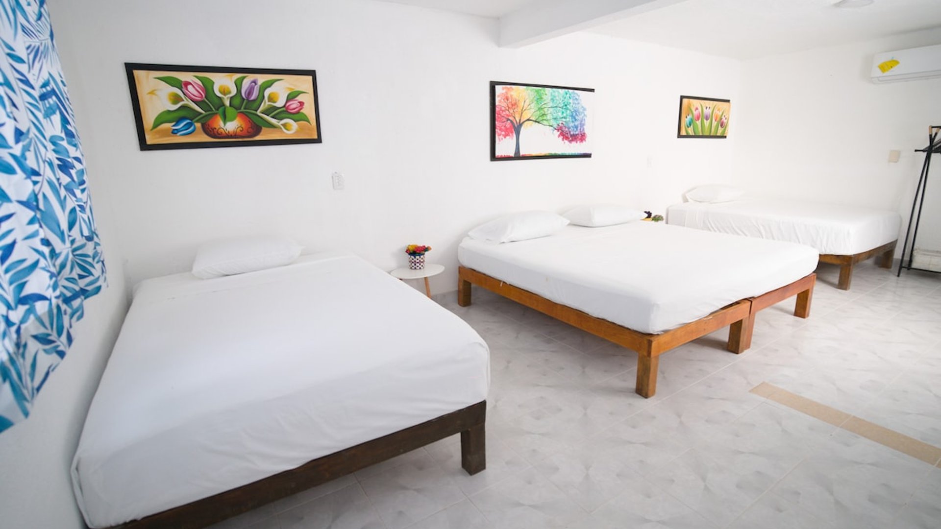 Recámara amplia con tres camas de madera, ropa de cama blanca y cuadros coloridos en paredes blancas