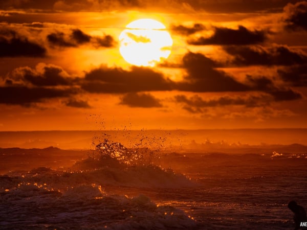 Atardecer intenso sobre el Pacífico con olas rompiendo en silueta bajo un sol ardiente entre nubes dramáticas