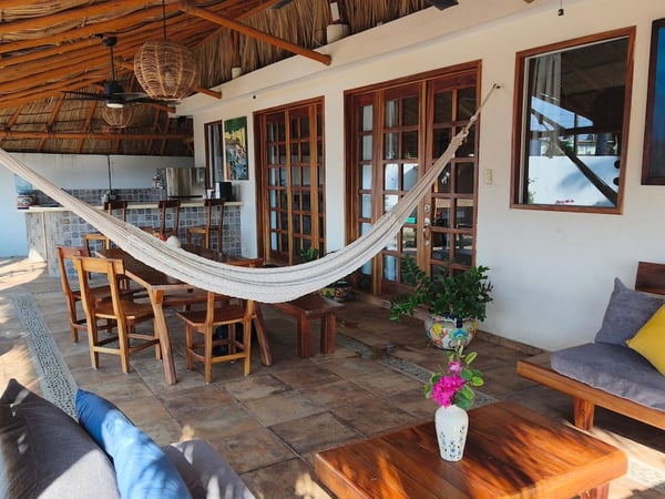 Terraza bajo palapa con hamaca, sala y comedor exterior rodeados de decoración artesanal mexicana