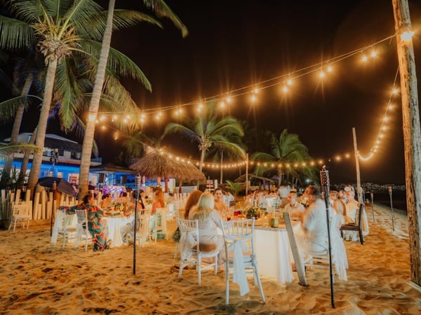 Banquete de boda nocturno en la playa con mesas blancas, luces colgantes y palmeras iluminadas