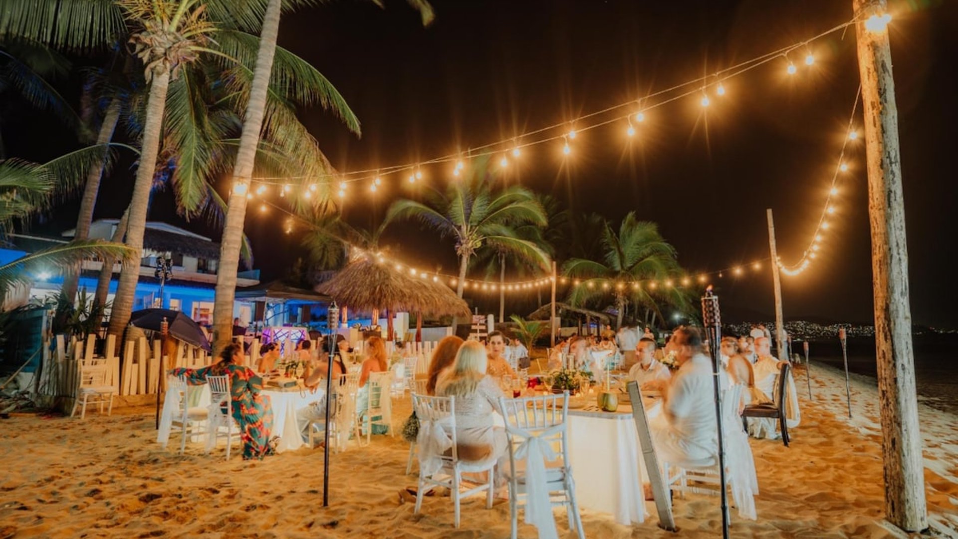 Banquete de boda nocturno en la playa con mesas blancas, luces colgantes y palmeras iluminadas