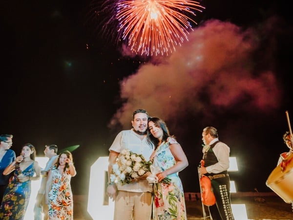 Pareja celebrando en la playa de noche con fuegos artificiales, mariachi y ramo de flores entre invitados