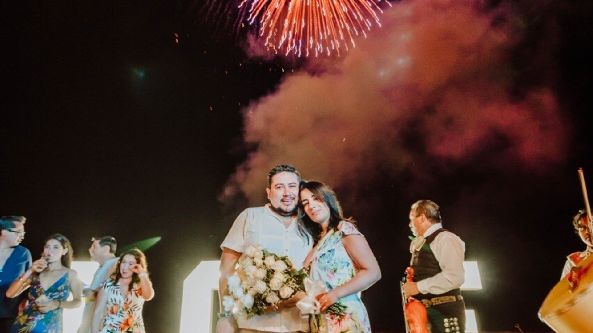 Pareja celebrando en la playa de noche con fuegos artificiales, mariachi y ramo de flores entre invitados