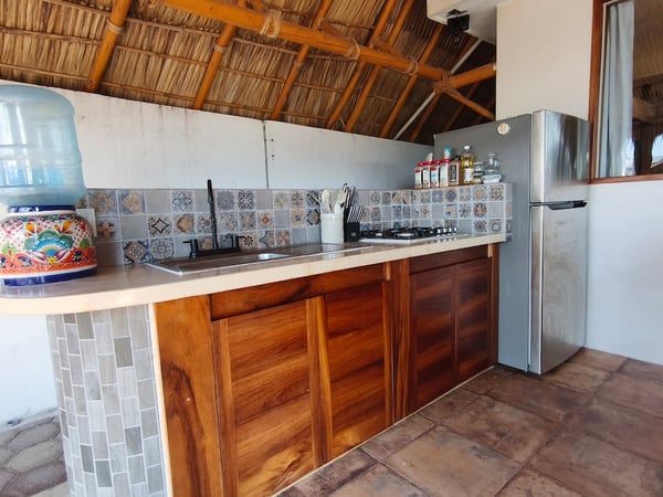 Cocina bajo palapa con gabinetes de madera, salpicadera de talavera, fregadero y refrigerador de acero inoxidable