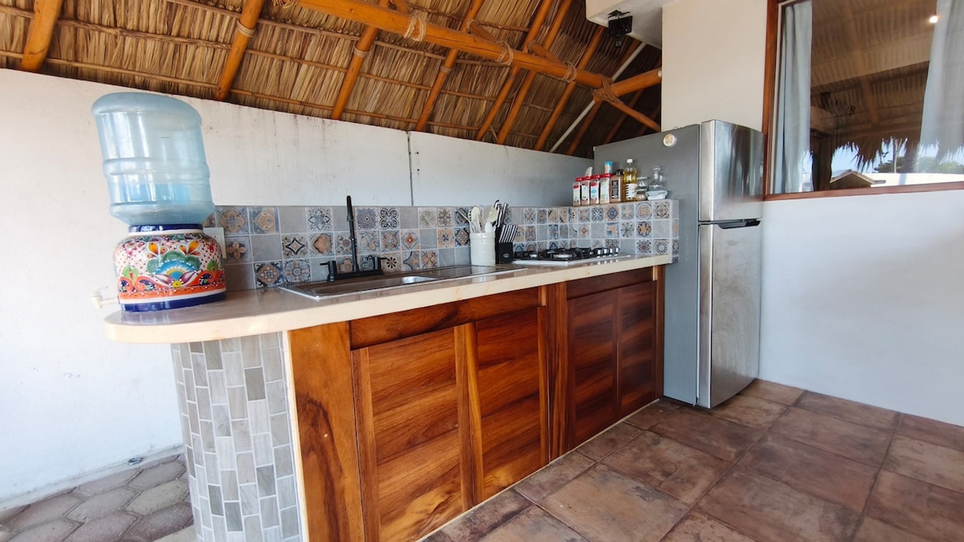 Cocina bajo palapa con gabinetes de madera, salpicadera de talavera, fregadero y refrigerador de acero inoxidable