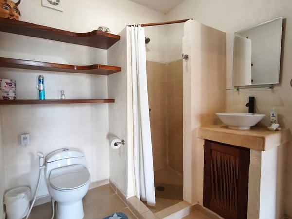 Baño completo con lavabo sobre mueble de cantera, regadera con cortina blanca y repisas de madera