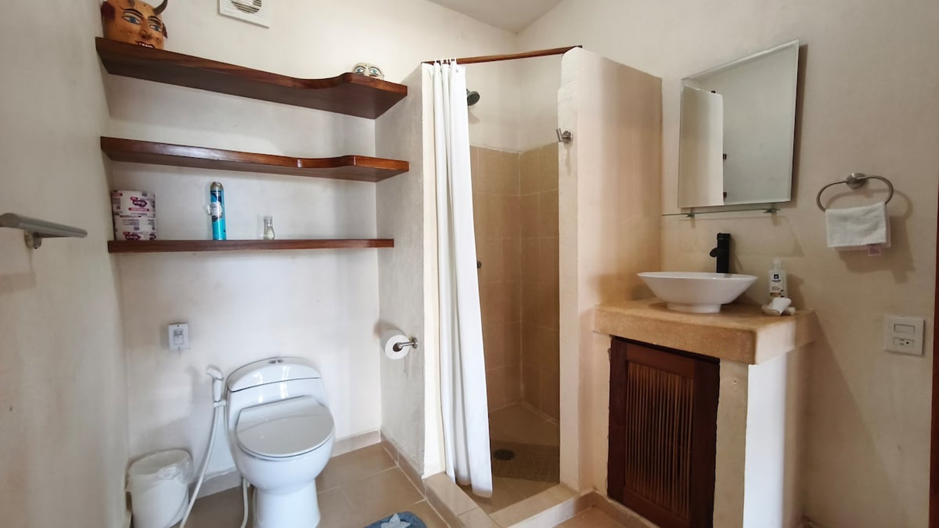 Baño completo con lavabo sobre mueble de cantera, regadera con cortina blanca y repisas de madera