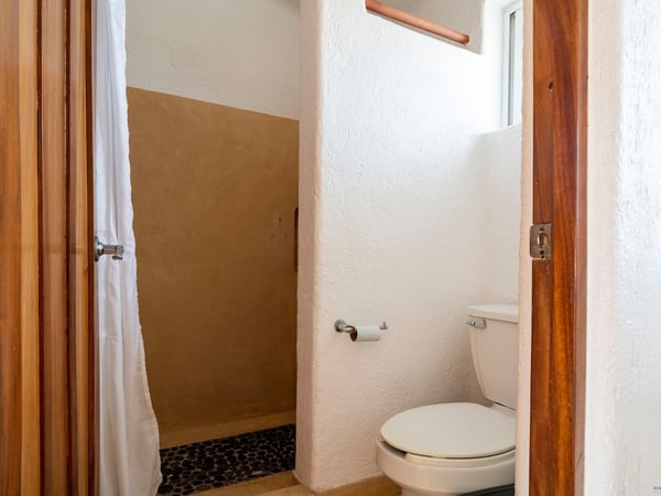 Baño con inodoro, ventana pequeña y acceso a regadera con cortina blanca y piso de piedra