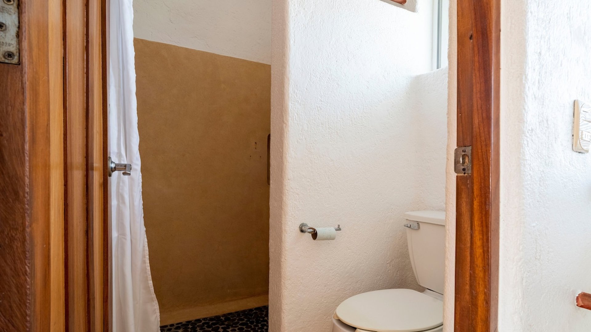 Baño con inodoro, ventana pequeña y acceso a regadera con cortina blanca y piso de piedra