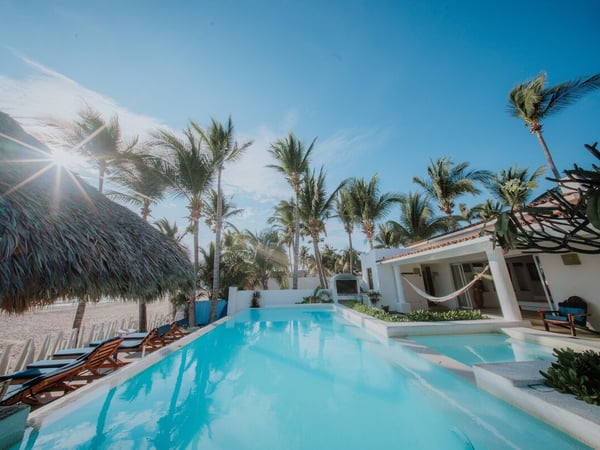 Alberca frente a la playa rodeada de palmeras, palapa y villa blanca con hamaca bajo cielo azul despejado