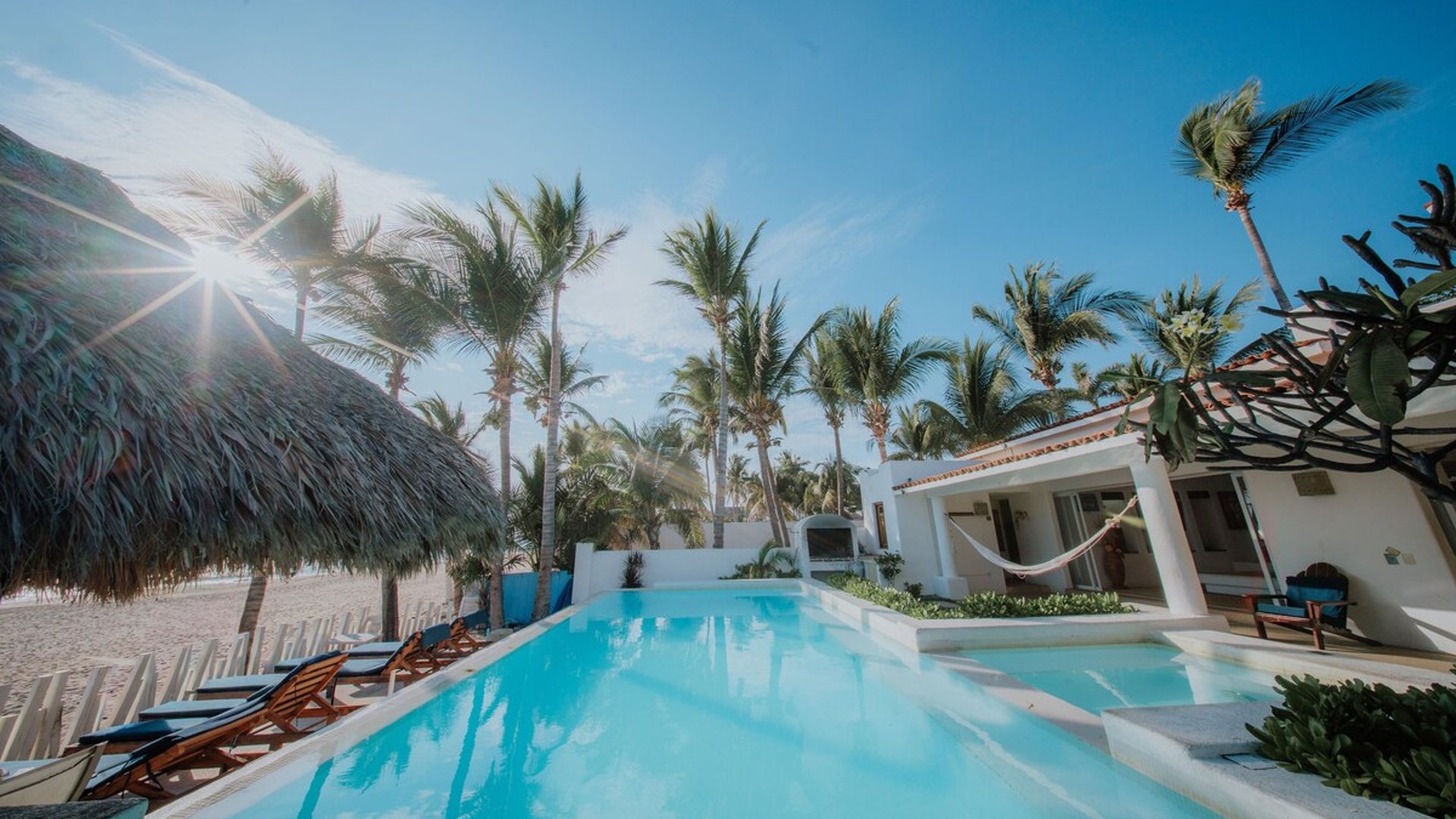 Alberca frente a la playa rodeada de palmeras, palapa y villa blanca con hamaca bajo cielo azul despejado