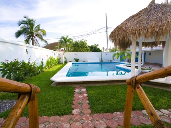 Jardín con alberca rectangular, palapa, pasto verde y palmeras en área exterior de la propiedad