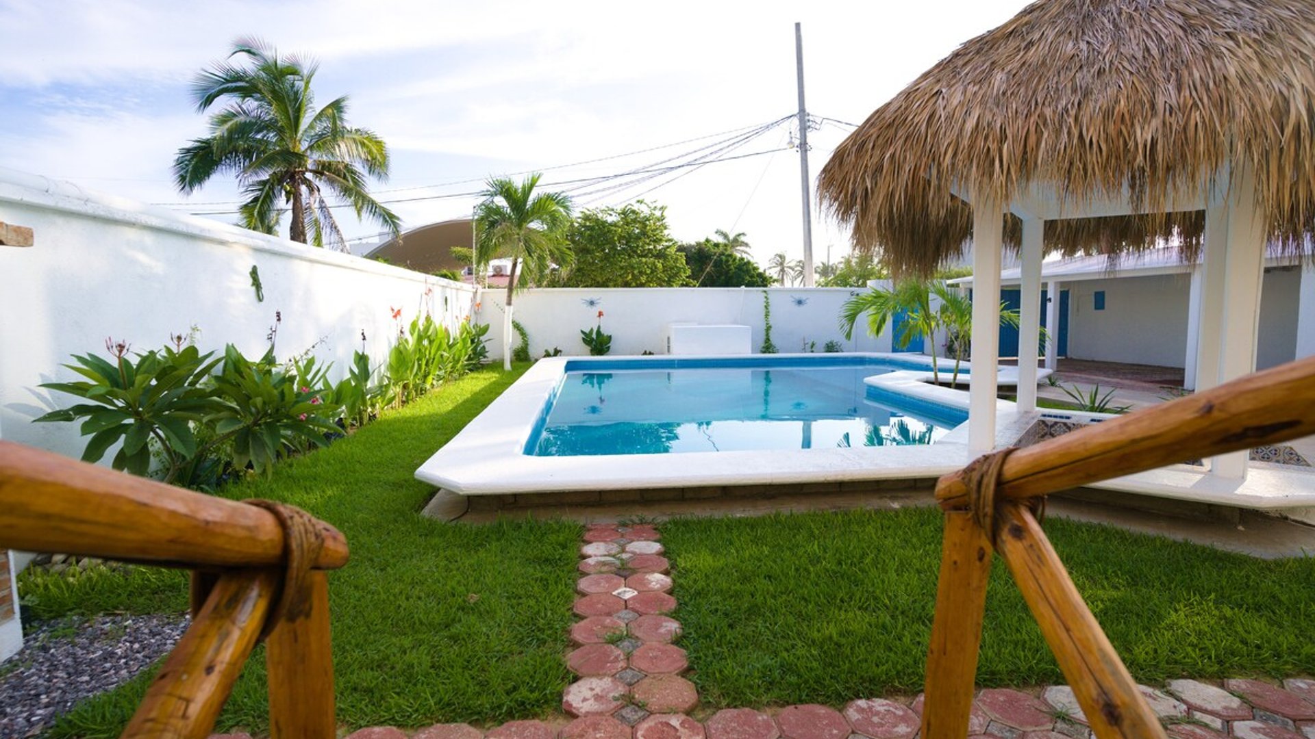 Jardín con alberca rectangular, palapa, pasto verde y palmeras en área exterior de la propiedad
