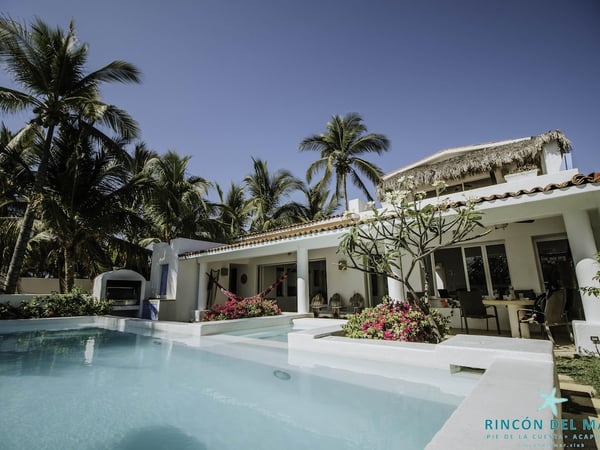 Villa blanca con alberca privada, hamaca, buganvilias y palmeras tropicales bajo cielo azul despejado