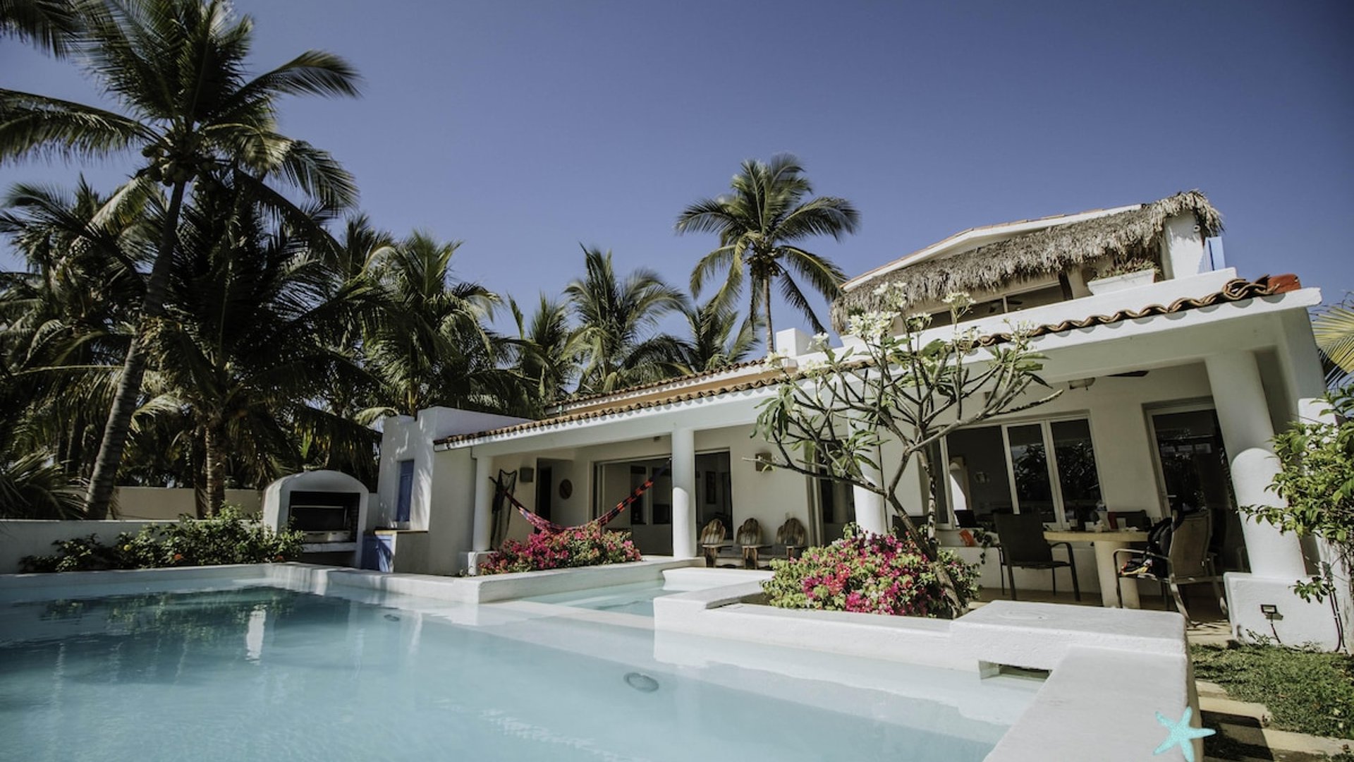 Villa blanca con alberca privada, hamaca, buganvilias y palmeras tropicales bajo cielo azul despejado