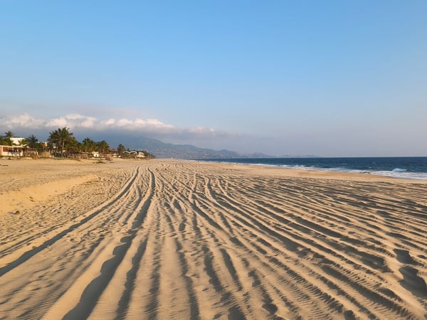Amplia playa de arena con ondulaciones al amanecer, mar azul y montañas al fondo con luz dorada