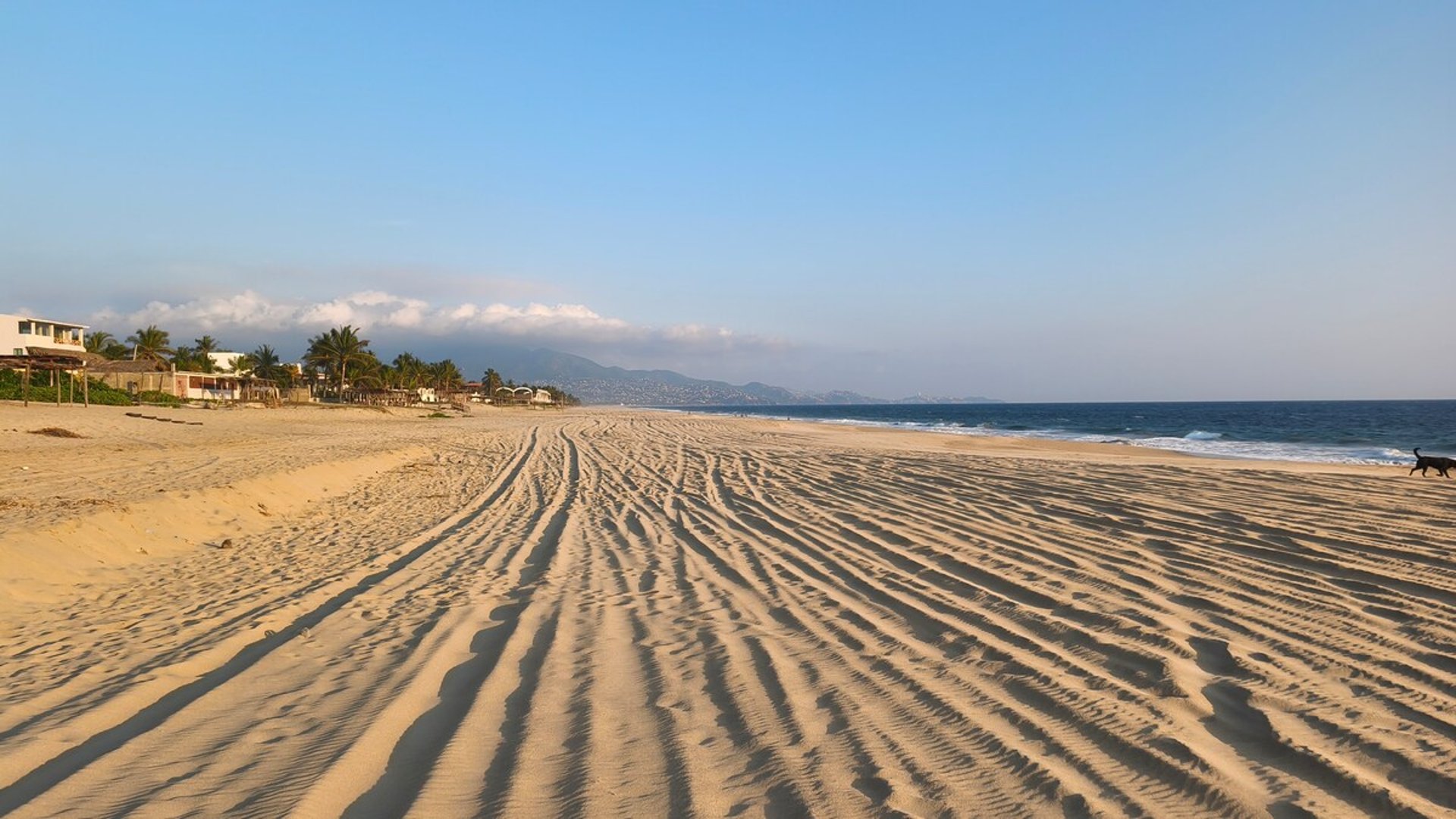 Amplia playa de arena con ondulaciones al amanecer, mar azul y montañas al fondo con luz dorada