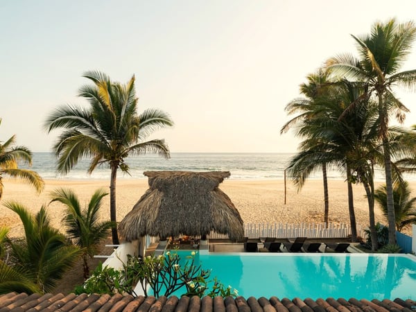 Alberca infinita con palapa de playa, palmeras y mar al fondo bajo luz dorada de atardecer
