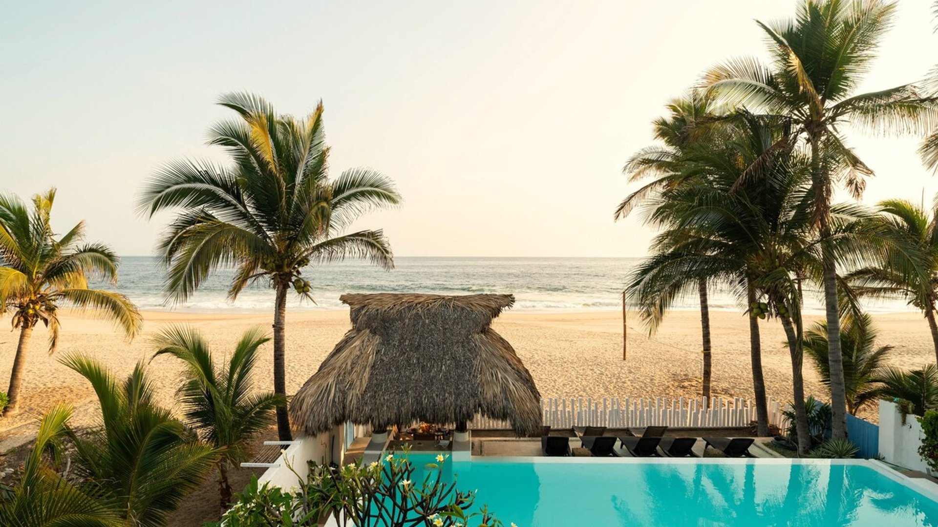 Alberca infinita con palapa de playa, palmeras y mar al fondo bajo luz dorada de atardecer