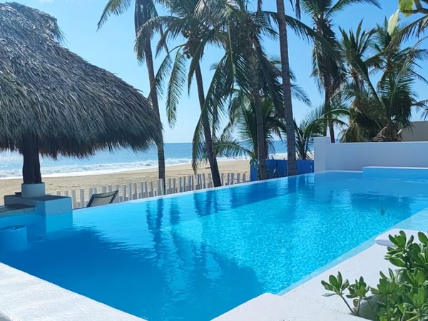 Alberca infinity de aguas cristalinas con palapa y vista directa a la playa y el mar bajo cielo despejado