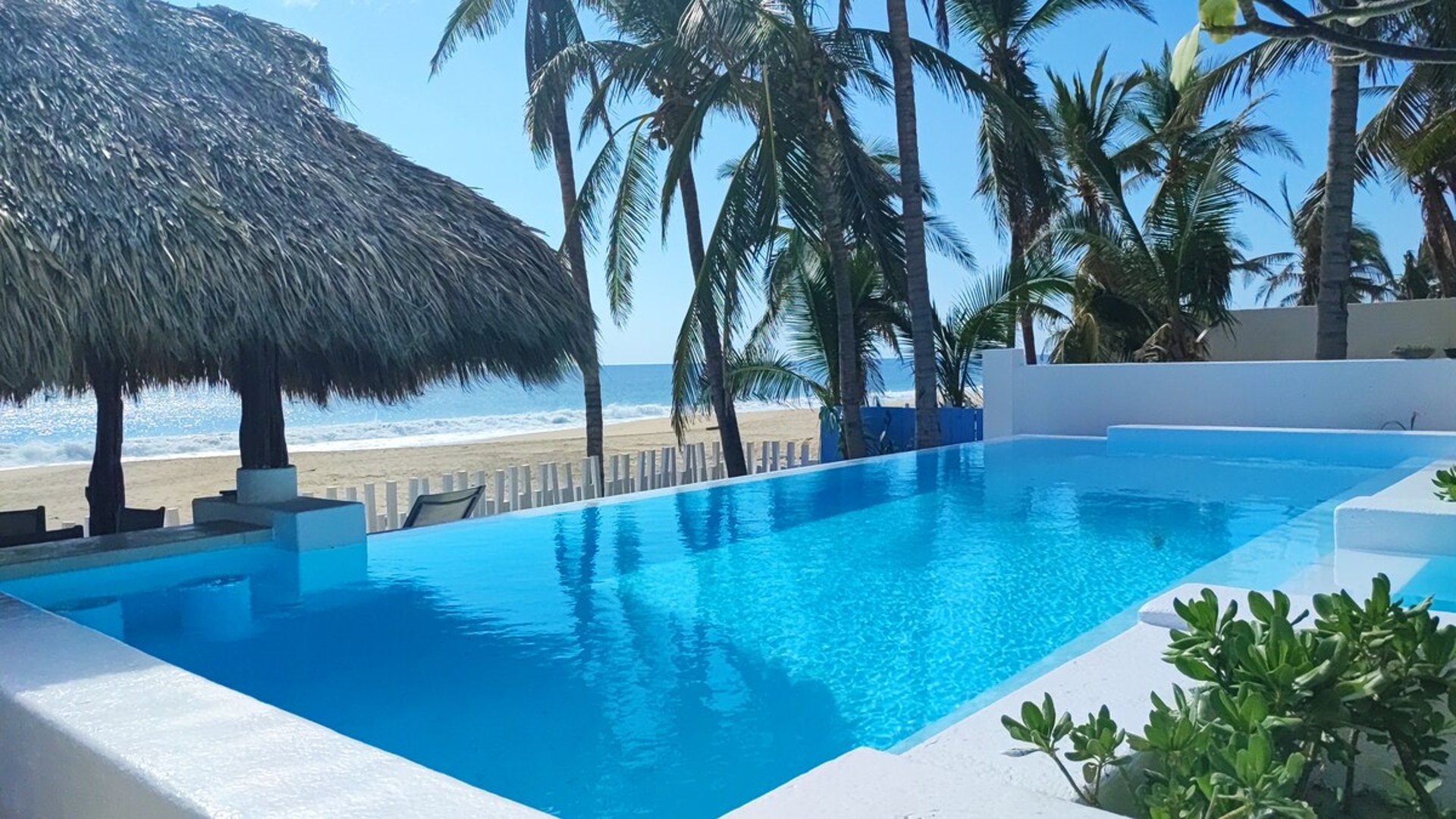 Alberca infinity de aguas cristalinas con palapa y vista directa a la playa y el mar bajo cielo despejado