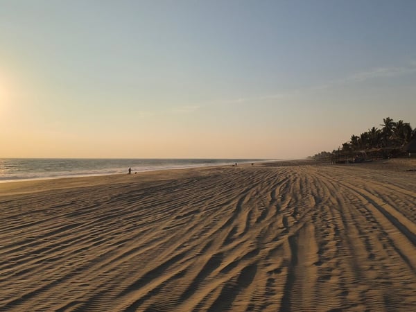 Amplia playa de arena con huellas al atardecer, mar tranquilo y palmeras al fondo bajo luz dorada